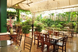 Un jardín interior con mesas y sillas de madera, rodeado de plantas diversas y una valla de bambú.