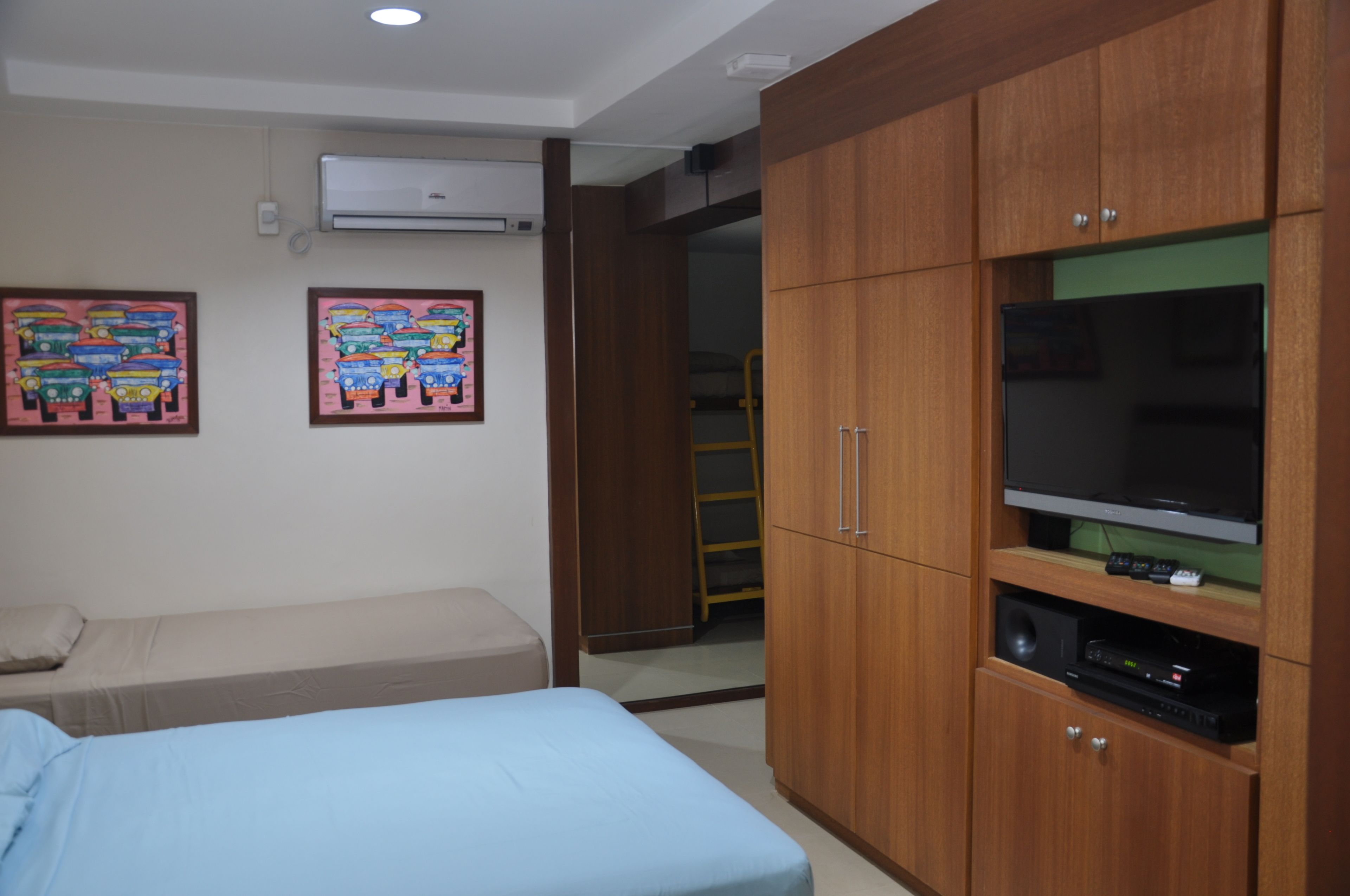 A bedroom with two single beds, a wardrobe, a television, and an air conditioning unit.