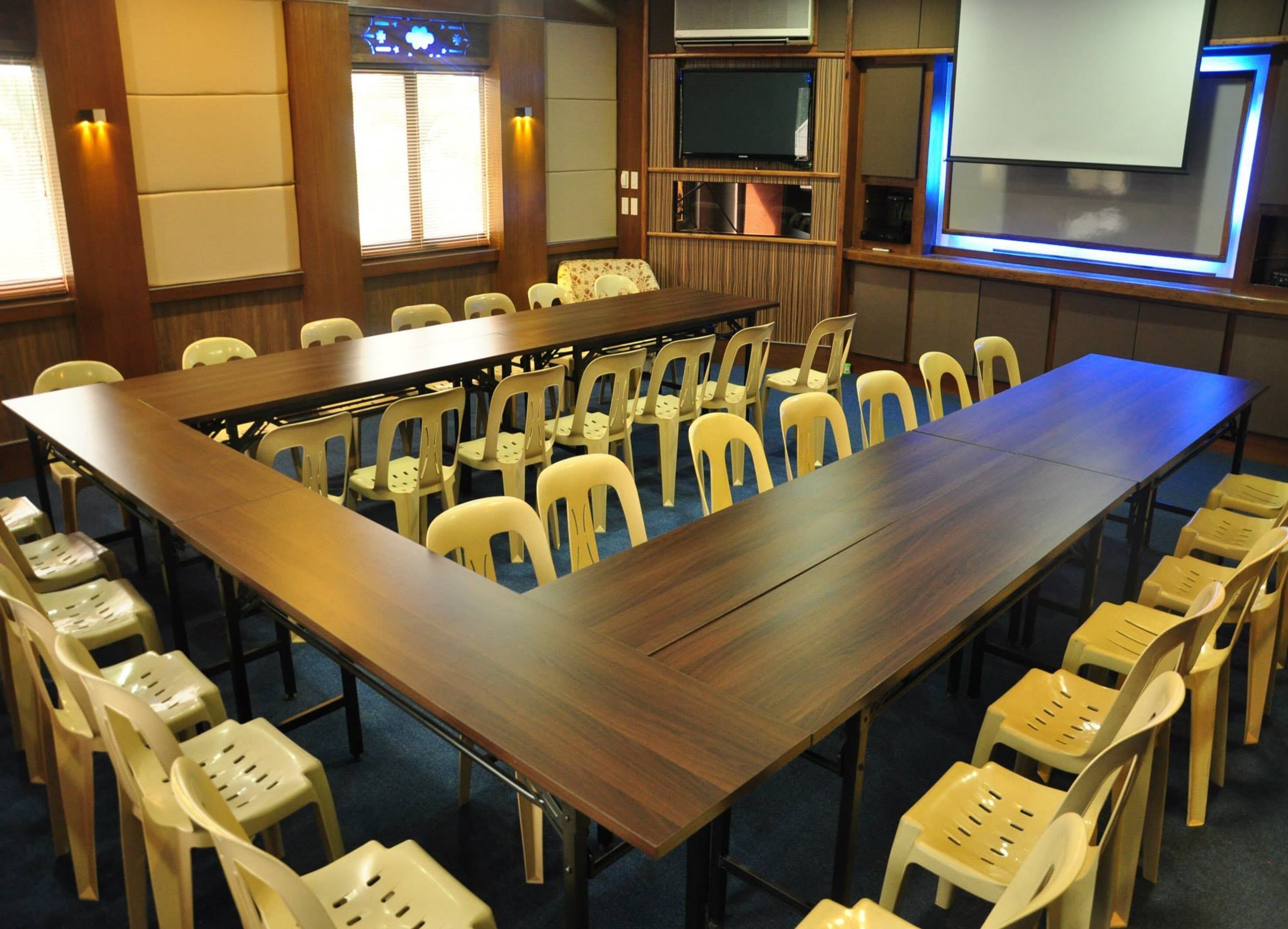 Uma sala de conferências com uma longa mesa de madeira rodeada por cadeiras de plástico, uma televisão na parede e um projetor.