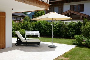 A covered outdoor seating area with a lounge chair and umbrella, a white bench, and a view of a mountain and residential buildings in the background. - Bodenschneid Suite Garden View West - Bodenschneid Suites Rottach-Egern (Rottach-Egern)