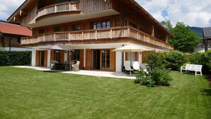 A two-story wooden house with a balcony, a patio with outdoor furniture, and a well-maintained lawn. - Bodenschneid Suite Garden View West - Bodenschneid Suites Rottach-Egern (Rottach-Egern)