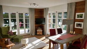 A living room with a TV, green and red chairs, a wooden table, and large windows. - Bodenschneid Suite Garden View West - Bodenschneid Suites Rottach-Egern (Rottach-Egern)
