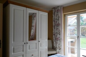 A room with a large white wardrobe, a chair, and a door leading to an outdoor area with a wicker table. - Bodenschneid Suite Garden View West - Bodenschneid Suites Rottach-Egern (Rottach-Egern)