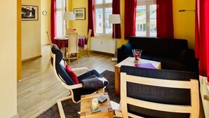 Ein modernes Wohnzimmer mit einer Couch, einem Sessel und einem kleinen Tisch. Ein Fenster mit roten Vorhängen und eine Lampe stehen auf dem Tisch.