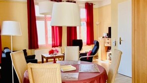 Ein Essbereich mit einem runden Tisch, der mit einer roten Tischdecke bedeckt ist und von Korbstühlen umgeben wird. Links steht eine Stehlampe, und auf dem Tisch steht eine Vase mit Blumen.