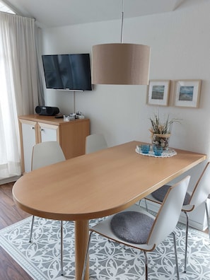 A modern hotel room with a wooden dining table, white chairs, a flat-screen TV, a radio, and framed artwork on the wall.