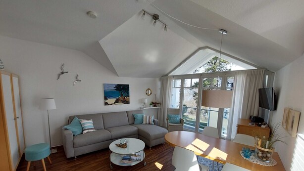 A modern living room with a grey sofa, a round coffee table, and a dining table with chairs. There is a large window providing natural light and a view of the outdoors.