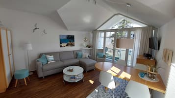 Ein modernes Wohnzimmer mit grauem Sofa, einem runden Couchtisch und einem Esstisch mit Stühlen. Ein großes Fenster lässt Tageslicht herein und bietet einen Ausblick ins Freie.