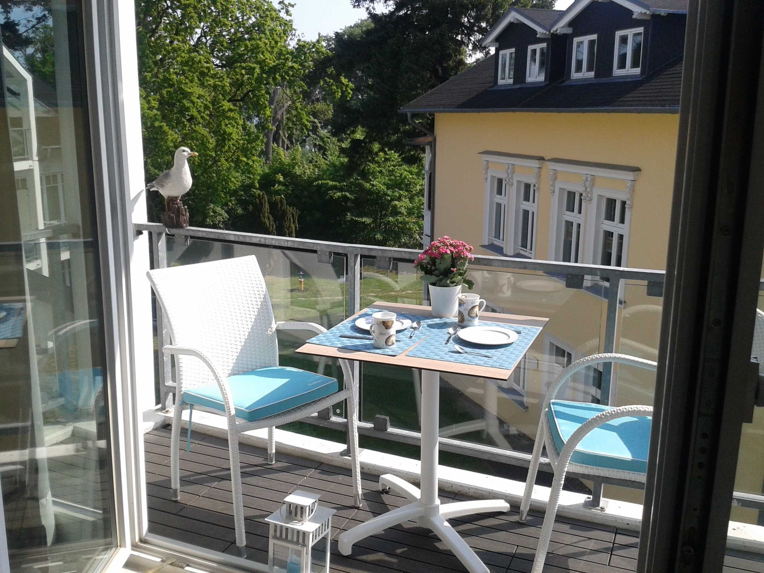Ein Balkon mit Tisch und Stühlen für zwei Personen, ein Vogel auf dem Geländer und ein gelbes Gebäude im Hintergrund.