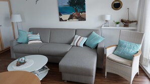 A living room with a grey sectional sofa, a wicker armchair, a small round table, and a painting of a beach scene.