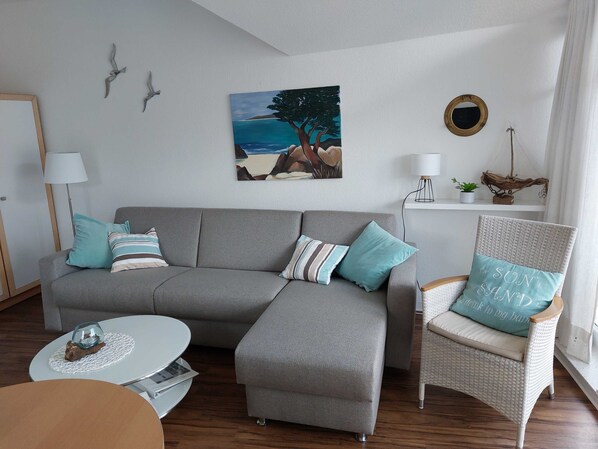 A living room with a grey sectional sofa, a wicker armchair, a small round table, and a painting of a beach scene.