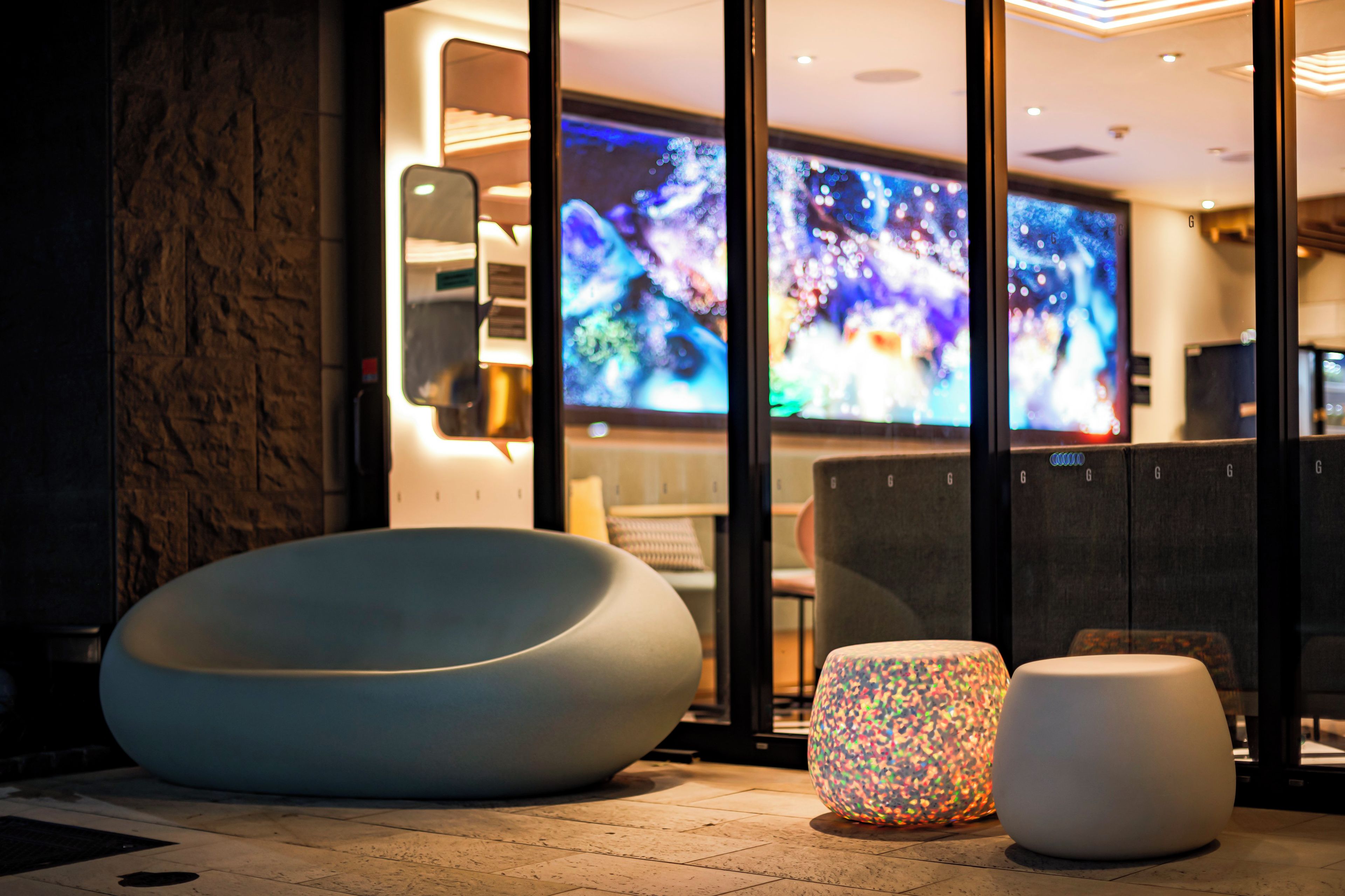 A modern interior with a large bean bag chair, a colorful illuminated stool, and a large digital screen displaying abstract art.