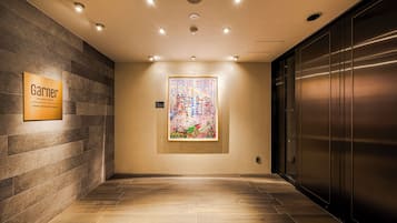 A hotel corridor with a modern elevator, a framed artwork on the wall, and a sign indicating the hotel's location.