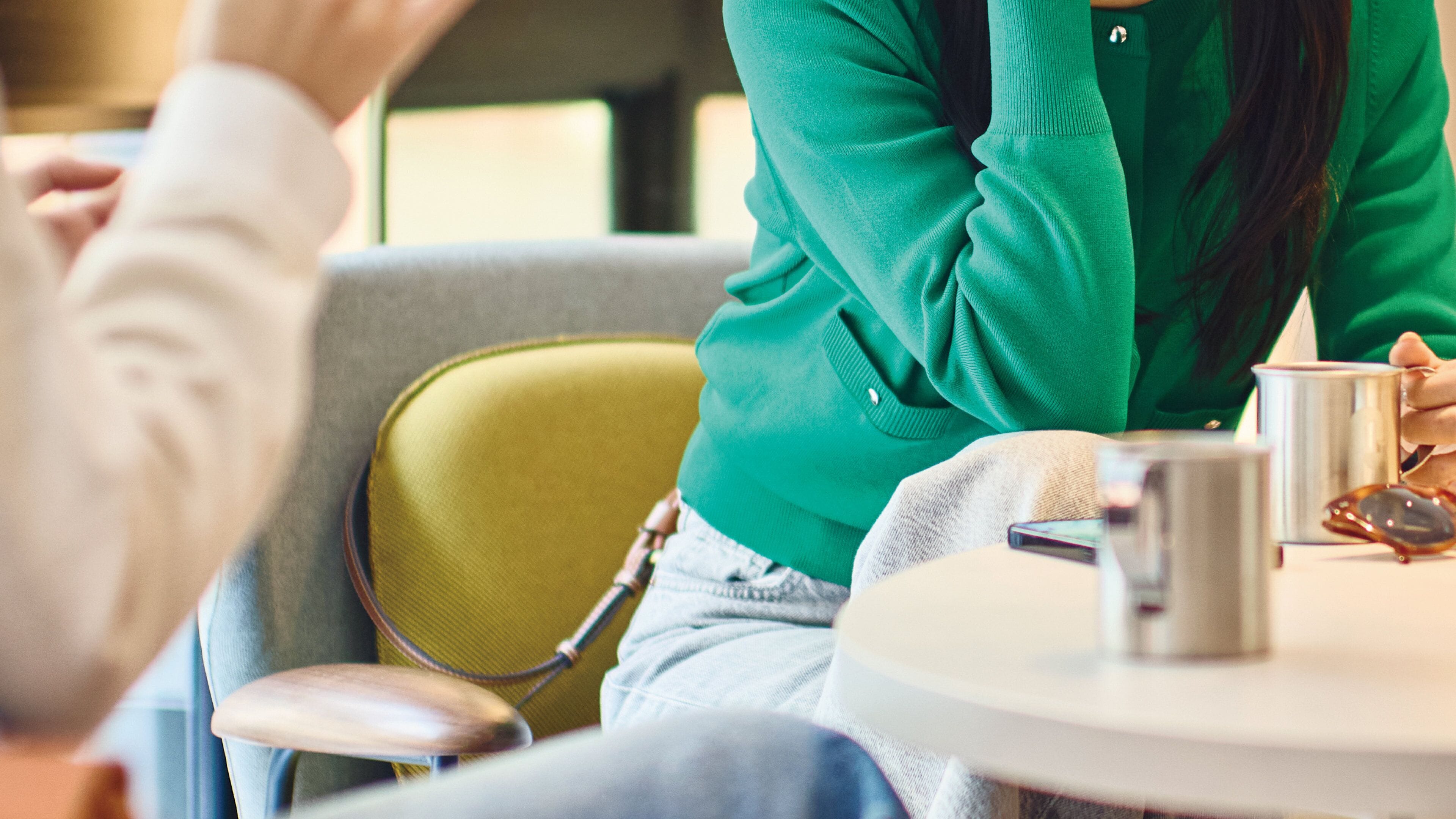 Una persona con suéter verde sentada a una mesa redonda, con un teléfono inteligente, una taza de metal y una cuchara.