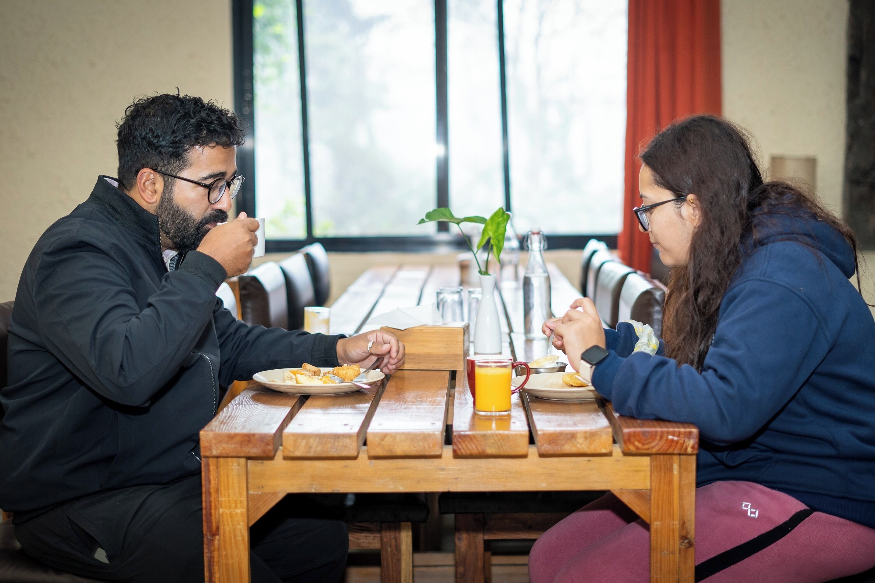 Two people sitting at a wooden table with a plant, a glass of orange juice, and a bottle of water.