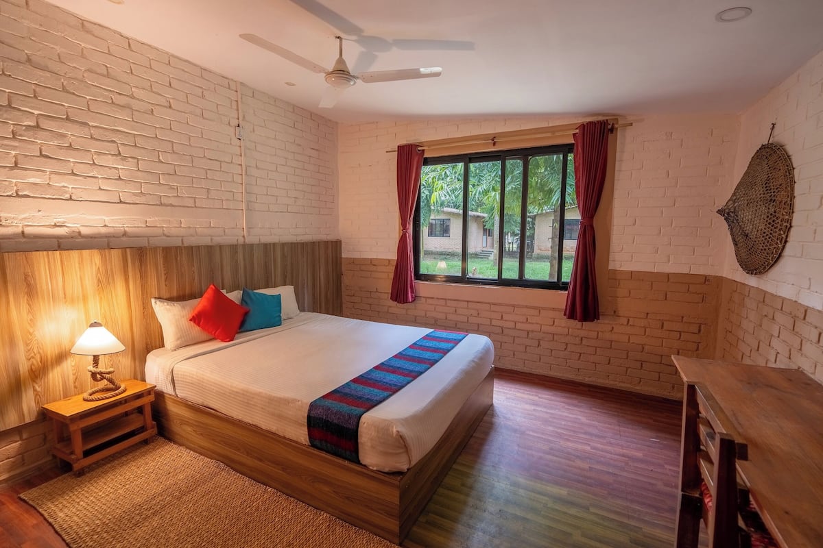 Un dormitorio con cabecera de madera, cama con manta colorida, mesita de noche con lámpara, ventana con cortinas y ventilador de techo.