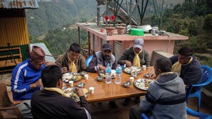 A group of people dining at a wooden table outdoors with a scenic view of hills in the background. - Mount Mahabharat Homestay (Dhunkharka)