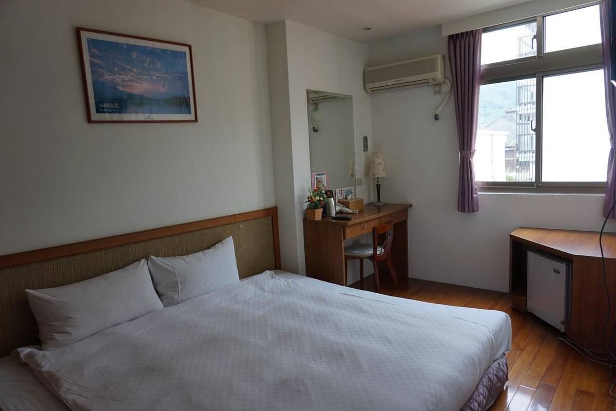 A hotel room with a bed, a desk, a chair, a small refrigerator, a window with curtains, and a wall-mounted air conditioner.