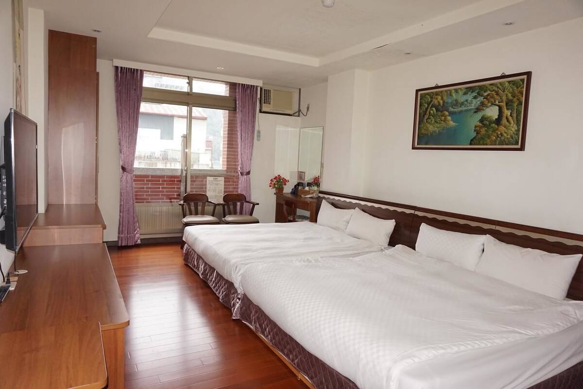 A hotel room with a large bed, a wooden desk, a chair, a window with curtains, a painting on the wall, and a radiator.