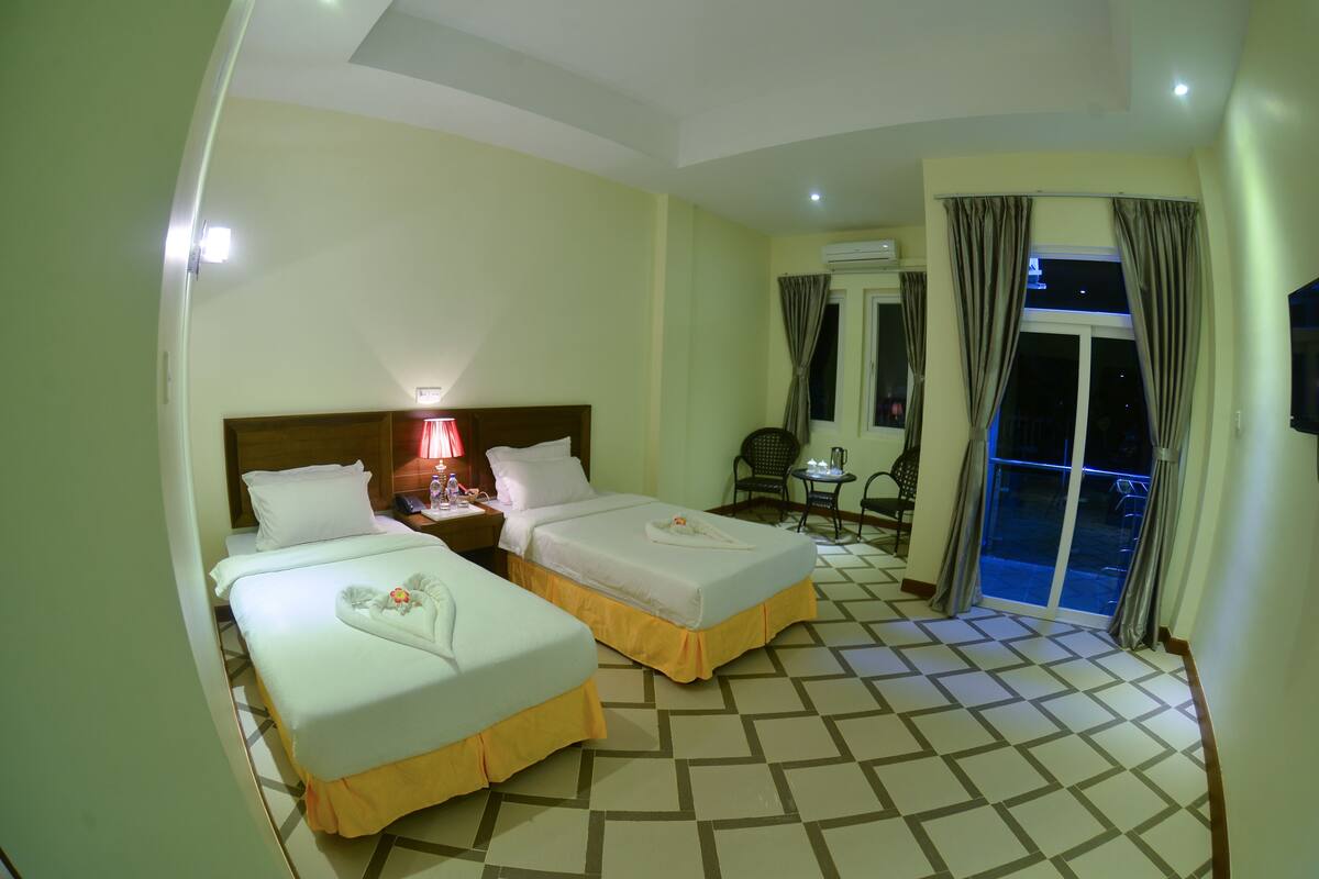 A hotel room with two beds, a small table with chairs, a balcony with curtains, and a tiled floor.