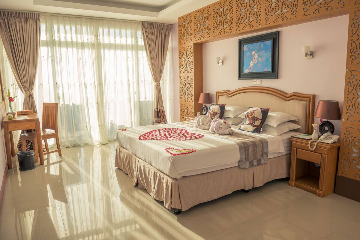 A hotel room with a large bed, a desk, a chair, a bedside table with a lamp, and a decorative wall panel.