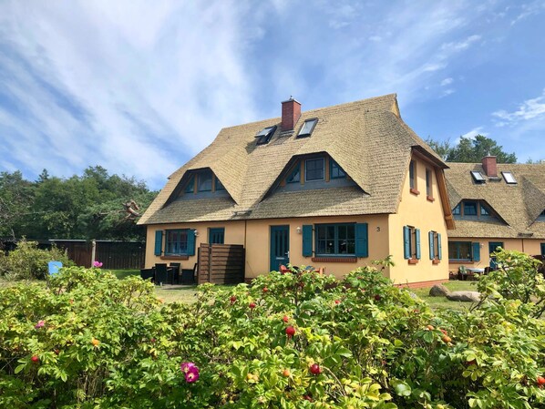 Ein traditionelles Haus mit einem Strohdach, zahlreichen Fenstern und einem Garten mit Obststräuchern.