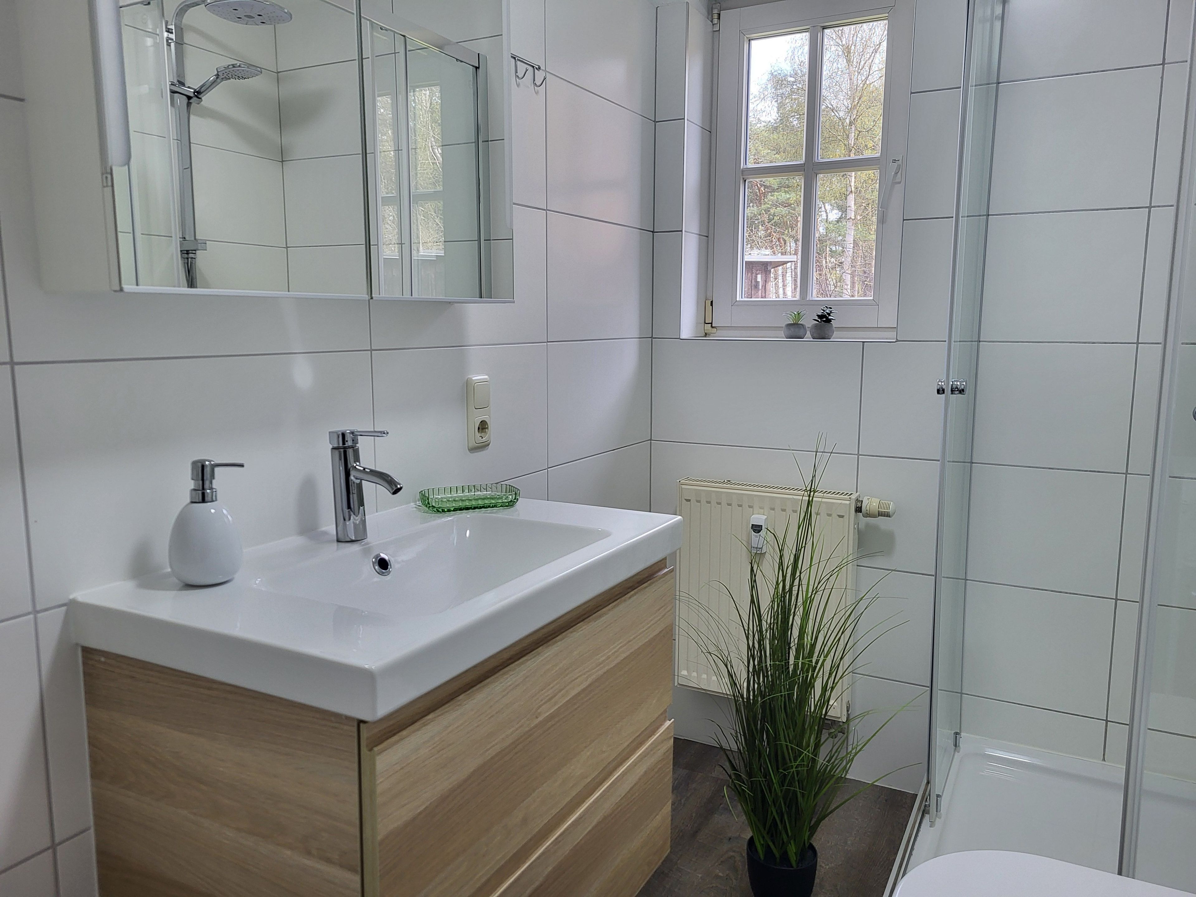 Ein modernes Badezimmer mit einem weißen Waschbecken, einem Spiegel, einer Dusche und einem Fenster.