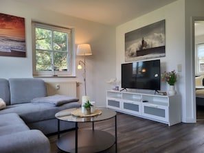 A modern living room with a grey sofa, a round coffee table, a TV on a white entertainment unit, a lamp, and a window with a view of trees. - Vacation farm at Fischland Flügge (Wustrow)