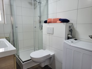 A bathroom with a glass-enclosed shower, a white toilet, and a washing machine. - Vacation farm at Fischland Flügge (Wustrow)