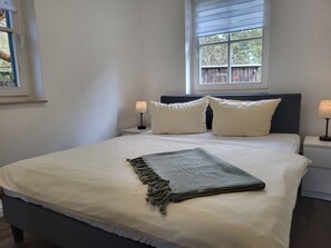 Ein ordentlich bezogenes Bett mit weißer Decke und zwei Kissen, eine Nachttischlampe, ein Fenster mit Jalousien und Blick auf Bäume.