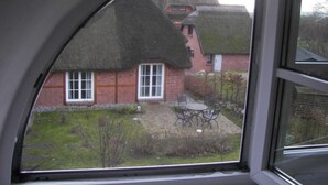 A view through a window of a house with a thatched roof, brick walls, and a small garden with a table and chairs.