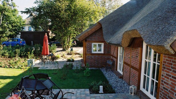 A thatched-roof house with a brick exterior, a paved patio with outdoor furniture, and a garden area with a lawn and trees.