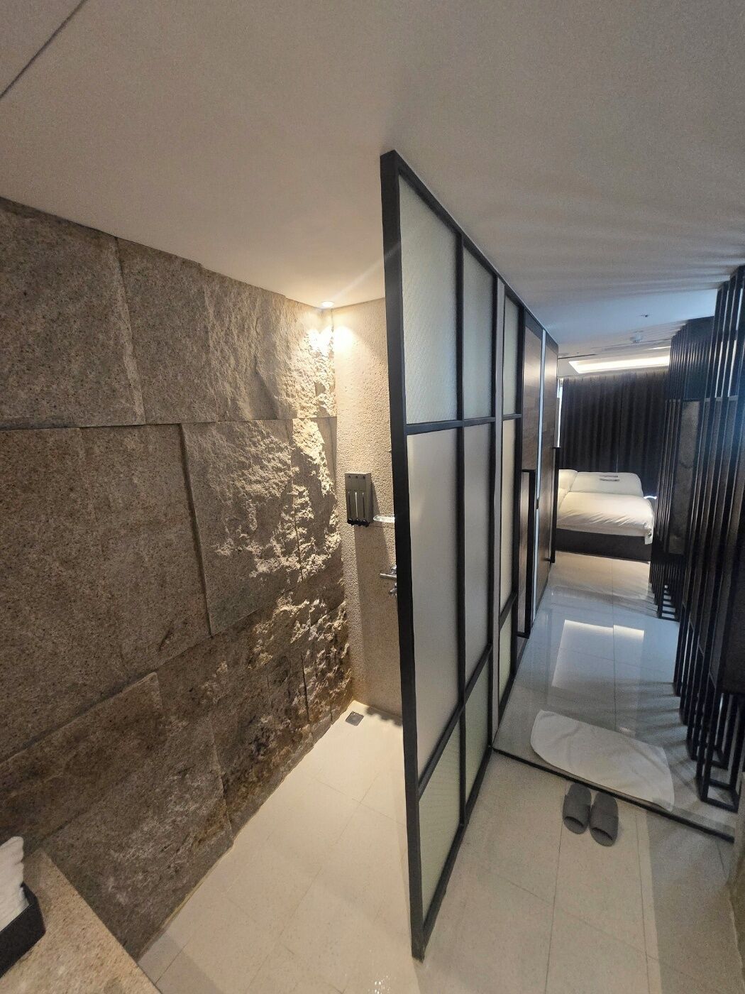 A modern bathroom with a stone wall, a glass partition, and a bed with a white mattress.