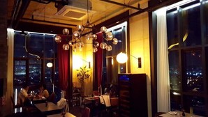Un restaurante con mesas y sillas, una araña y amplias ventanas con vistas a la ciudad.