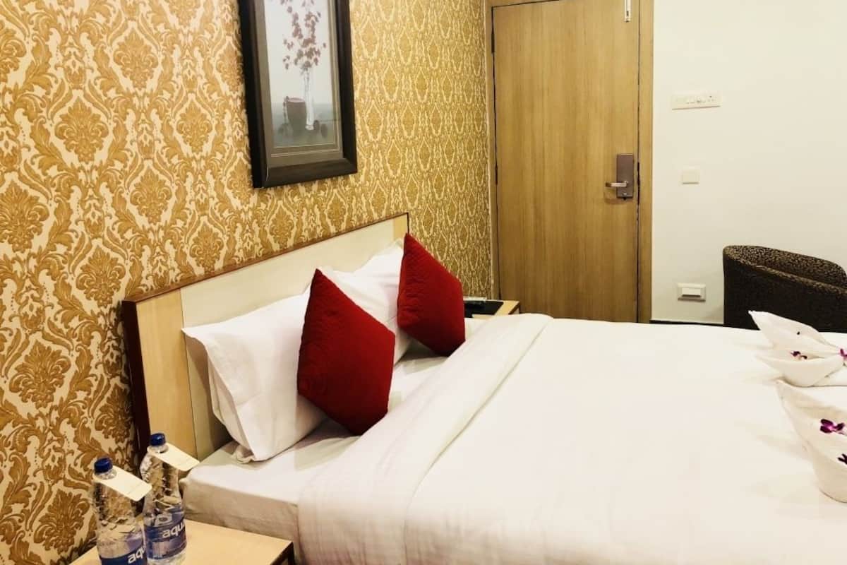 Habitación de hotel con papel tapiz estampado, un cuadro en la pared, una puerta de madera y una cama con ropa blanca y almohadas rojas.