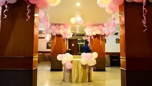 Un vestíbulo de hotel con un mostrador de recepción central, una araña y globos de color rosa y blanco decorando el espacio.