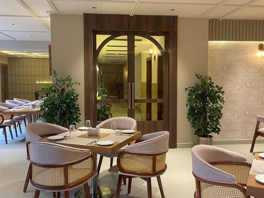 Un comedor moderno con mesas de madera y sillas de mimbre, un espejo grande y plantas en macetas.