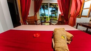 A hotel room with a bed, red curtains, a view of the beach, and a tropical garden.