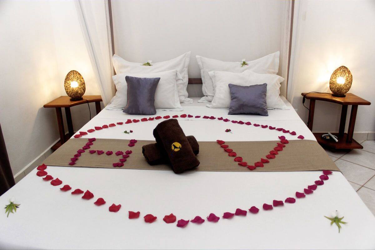 Una cama bien hecha con pétalos de rosa formando un corazón, una toalla marrón con un logo amarillo y dos lámparas de noche sobre mesitas de madera.