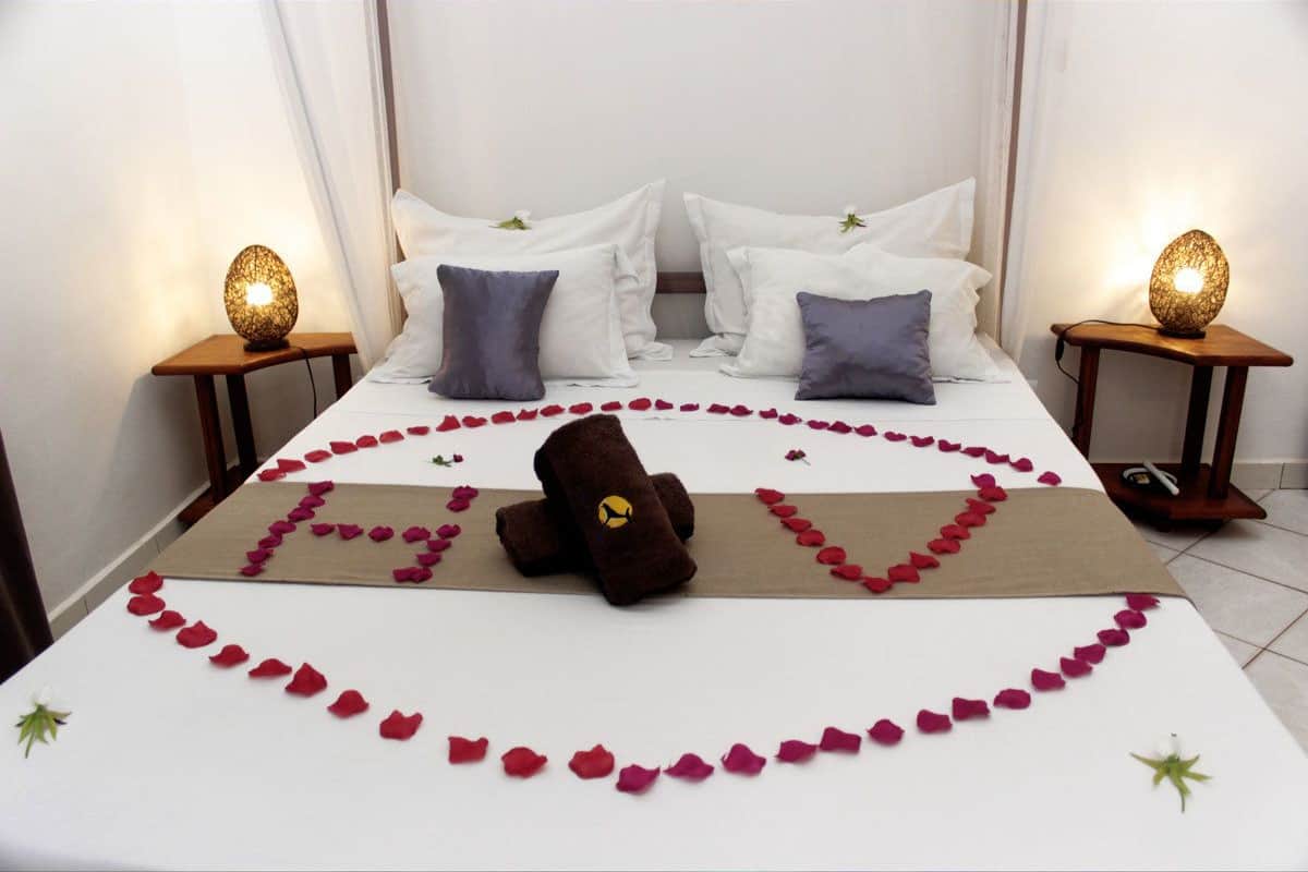 Una cama bien hecha con pétalos de rosa formando un corazón, una toalla marrón con un logo amarillo y dos lámparas de noche sobre mesitas de madera.