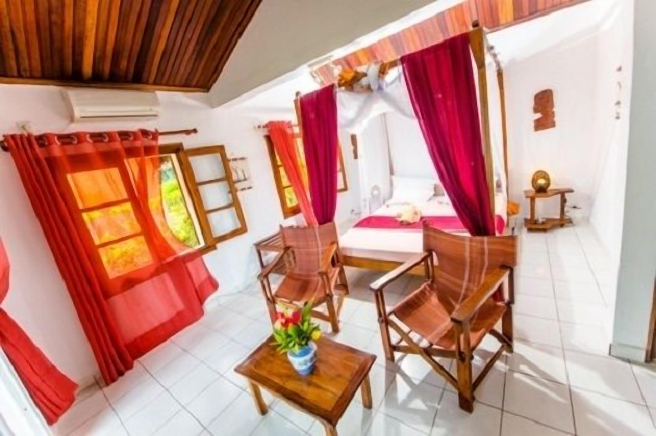 A room with a bed, two wooden chairs, a small table with a plant, and red curtains.