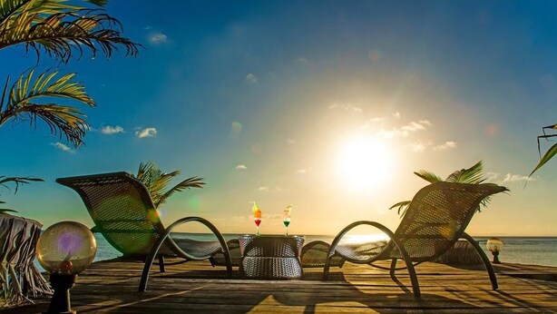 A wooden deck with two wicker chairs and a small table, overlooking the ocean at sunset.