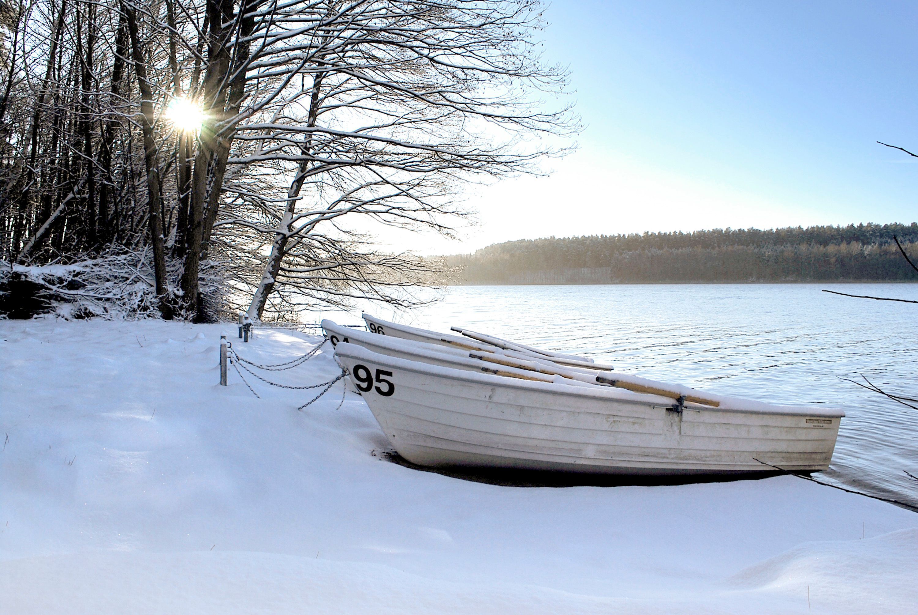 Ein mit Schnee bedecktes Boot, das mit einer Kette an einem Steg festgemacht ist, mit blauem Himmel und einem Wald im Hintergrund.