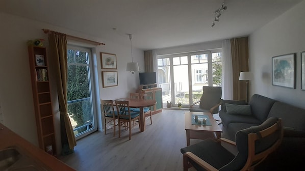 A spacious living area with a sofa, a dining table with chairs, a TV, and a balcony with a view of the outside.