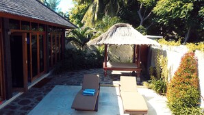 Un espace extérieur aménagé avec un toit de chaume, comprenant deux chaises longues et une table, entouré de plantes tropicales et bordé d’une terrasse pavée de pierres.