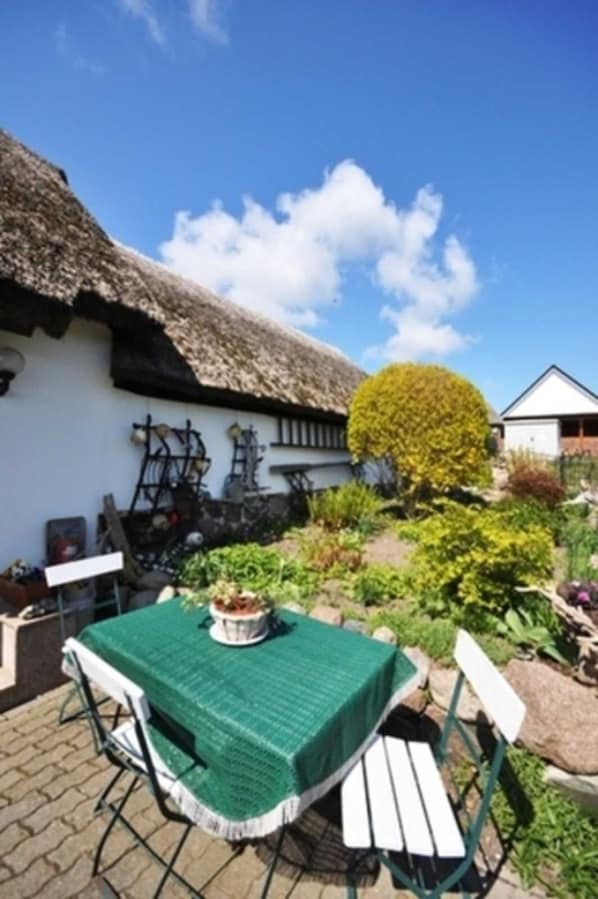 Une petite maison au toit de chaume, avec une table et des chaises vertes disposées à l’extérieur.