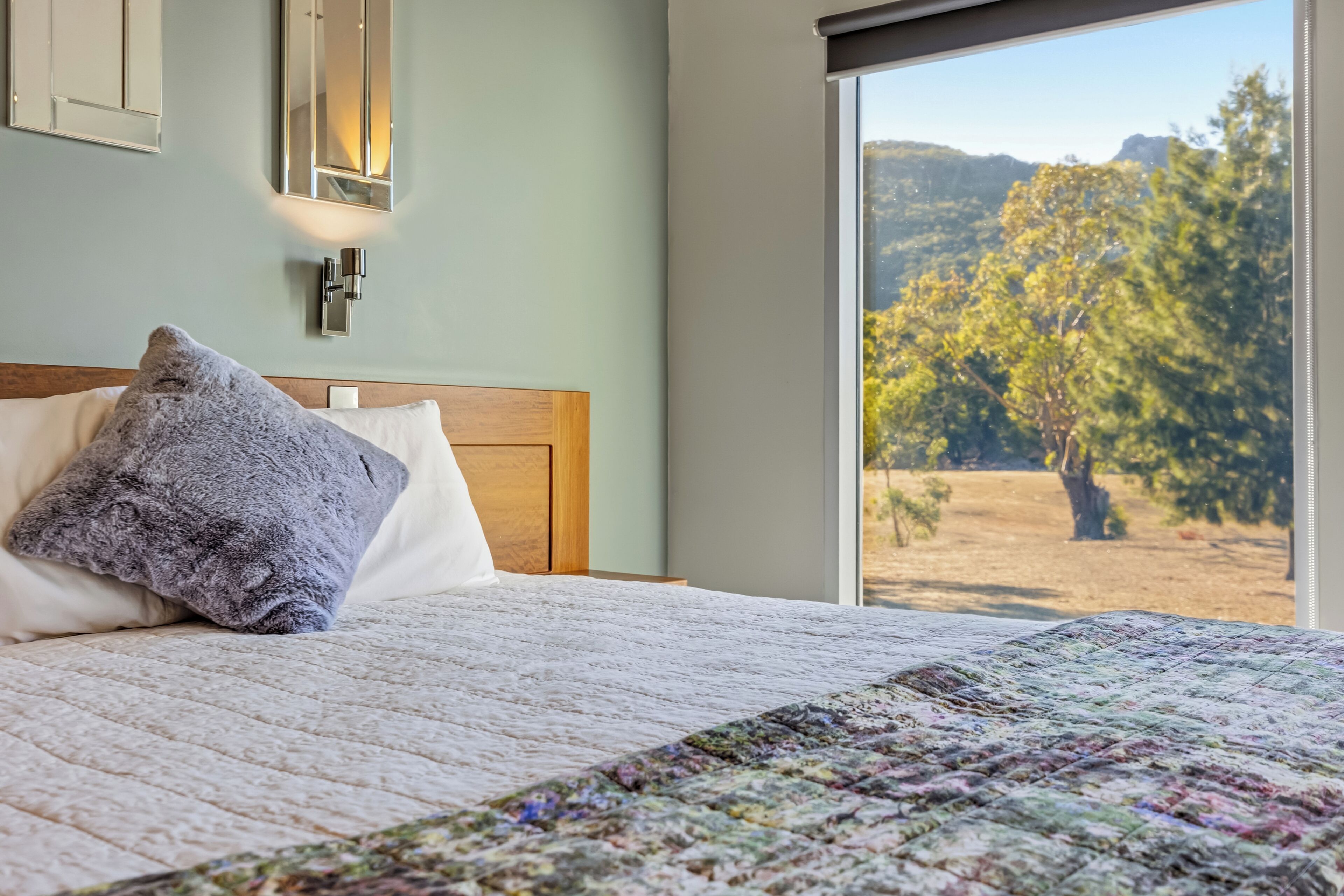 Uma cama com colcha, travesseiros, uma mesa de cabeceira, uma luminária fixada na parede e uma janela com vista para árvores e colinas.