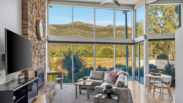 A spacious living room with a large TV, stone wall, and a view of the countryside. - Bushland (Halls Gap)