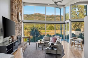 A spacious living room with a large TV, stone wall, and a view of the countryside. - Bushland (Halls Gap)