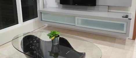 A modern living room with a glass coffee table, a flat-screen TV mounted on a white wall, and a sliding glass door with curtains.